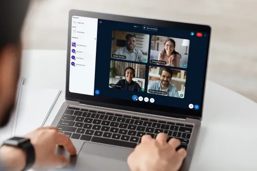 Person using a laptop showing a video conference interface with four participants in a grid, a participant list on the left, and meeting controls along the bottom.